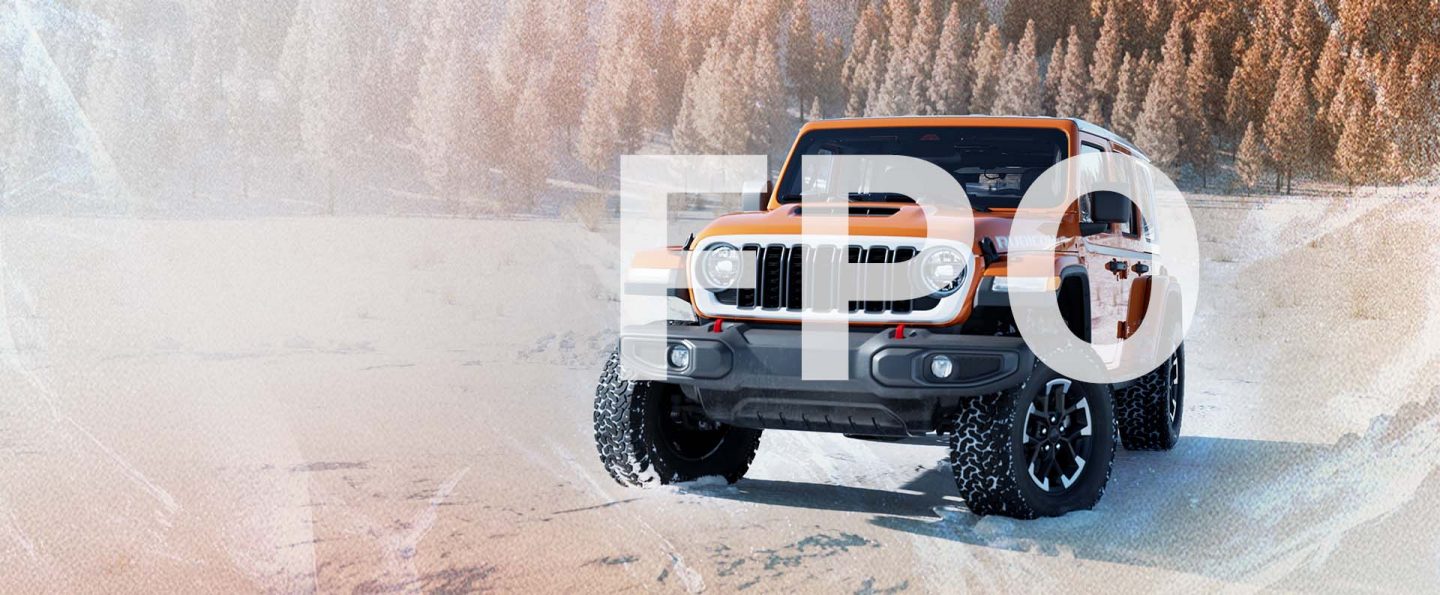 An orange 2026 Jeep Wrangler Rubicon Whitecap traveling down a snow-covered road with a forest of evergreens in the background.